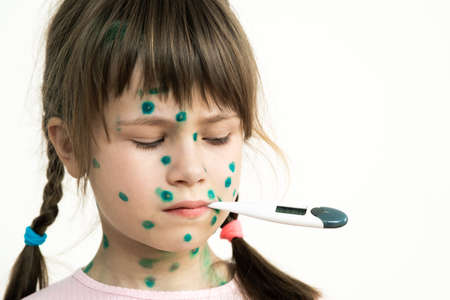 Child girl covered with green rashes on face ill with chickenpox, measles or rubella virus holding medical thermometer in her mouth having high temperature with fever.の写真素材