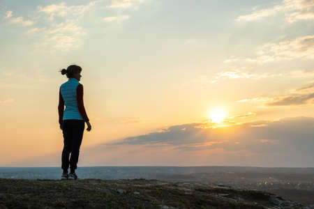 Silhouette of a woman hiker standing alone enjoying sunset outdoors. Female tourist on rural field...