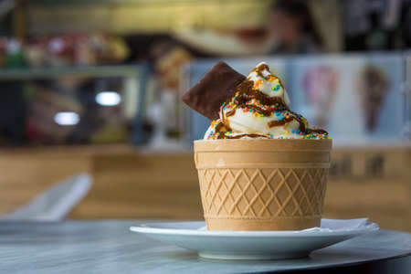 White plate with ice cream dessert in wafer cup with chocolate cookies and creative decoration topping on blurred colorful interior background.の写真素材