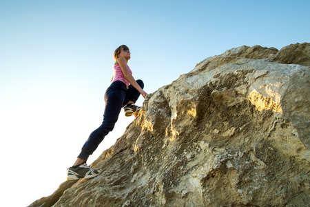 Woman hiker climbing steep big rock on a sunny day. Young female climber overcomes difficult...