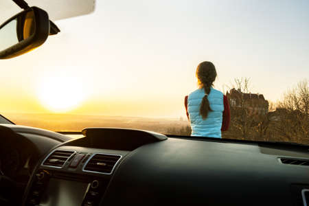 View from inside of a young woman standing near her car enjoying warm sunset view. Girl traveler...
