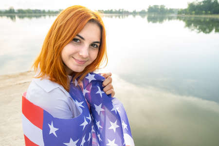 Portrait of happy smiling red haired girl with USA national flag on her shoulders. Young woman celebrating United States independence day.の写真素材