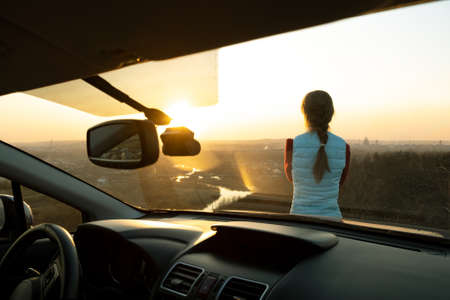 Young woman standing near her car enjoying warm sunset view. Girl traveler leaning on vehicle hood looking at evening horizon.の写真素材