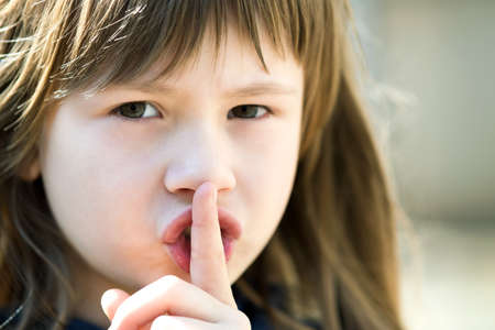 Portrait of pretty child girl with gray eyes and long hair holding point finger on her lips with hush sigh. Cute female kid on warm summer day outside.の写真素材