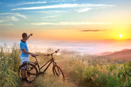 Man cyclist standing near his bicycle enjoying beautiful sunrise in mountains with white fog below.の写真素材