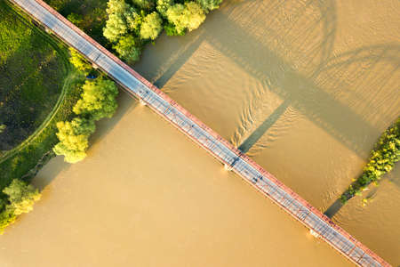 Aerial view of a narrow road bridge stretching over muddy wide river in green rural area.の写真素材