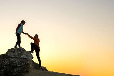 Man and woman hikers helping each other to climb a big stone at sunset in mountains. Couple climbing on a high rock in evening nature. Tourism, traveling and healthy lifestyle concept.の写真素材