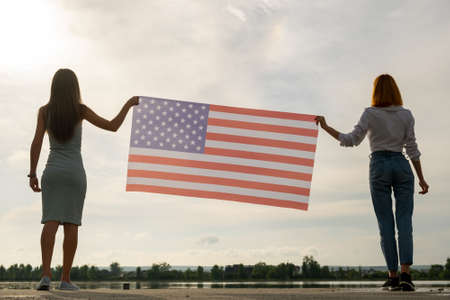 Silhouette of two young friends women holding USA national flag in their hands standing together...