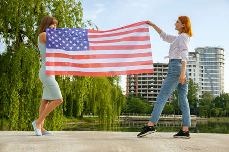 Two young friends women holding USA national flag in their hands standing together outdoors. Patriotic girls celebrating United States independence day.の写真素材