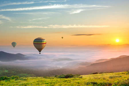 Big hot air baloons over idyllic landscape with green grass covered morning mountains with distant peaks and wide valley full of thick white cloudy fog.の写真素材