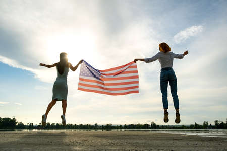 Two young friends women holding USA national flag jumping up together outdoors at sunset. Patriotic girls celebrating United States independence day.の写真素材