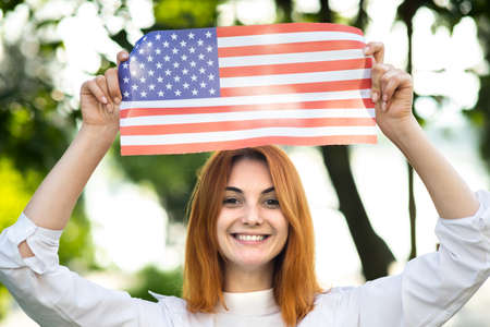 Portrait of happy young red haired woman holding USA national flag in her hands standing outdoors in summer park. Positive girl celebrating United States independence day.の写真素材