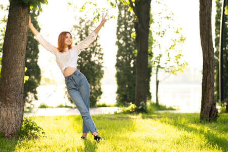 Young positive redhead woman in white blouse and blue jeans standing in summer park enjoying warm day.の写真素材