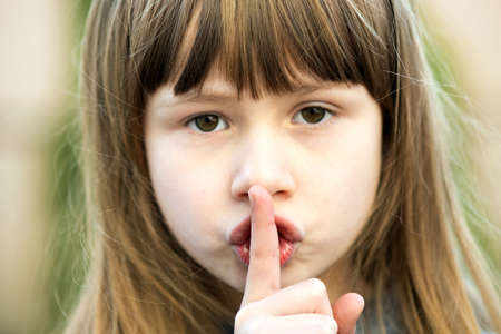 Portrait of pretty child girl with gray eyes and long fair hair holding point finger on her lips with hush sigh. Cute female kid on warm summer day outside.の写真素材