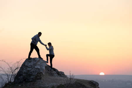 Man and woman hikers helping each other to climb stone at sunset in mountains. Couple climbing on high rock in evening nature. Tourism, traveling and healthy lifestyle concept.の写真素材