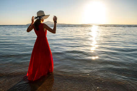 Young woman wearing long red dress and straw hat standing in sea water at the beach enjoying view of rising sun in early summer morning.の写真素材