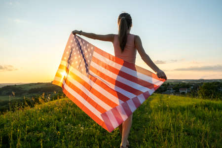 Happy young woman posing with USA national flag standing outdoors at sunset. Positive girl celebrating United States independence day. International day of democracy concept.の写真素材