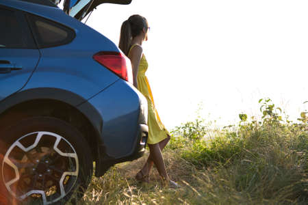 Happy young woman driver in yellow dress enjoying warm summer evening standing beside her car. Traveling and vacation concept.の写真素材