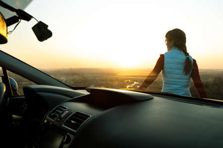 View from inside of a young woman standing near her car enjoying warm sunset view. Girl traveler...