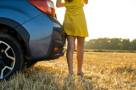 Back view of young woman driver slim legs in short yellow summer dress standing near her car on dry...
