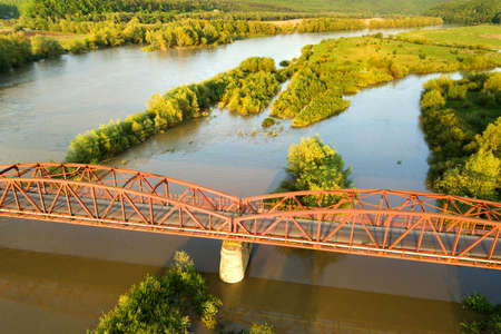 Aerial view of a narrow road bridge stretching over muddy wide river in green rural area.