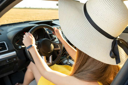 Driver woman in yellow dress and straw hat sitting behind steering wheel driving a car. Summer...