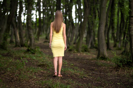 Back view of a slim woman in yellow dress standing in moody dark forest.