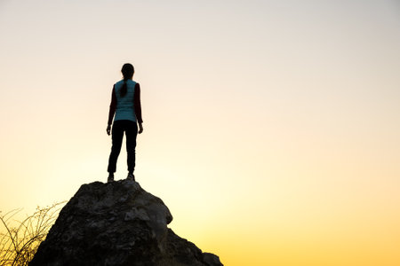 Silhouette of a woman hiker standing alone on big stone at sunset in mountains. Female tourist on high rock in evening nature. Tourism, traveling and healthy lifestyle concept.の写真素材