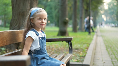 Small happy child girl sitting on a bench resting in summer park.の写真素材