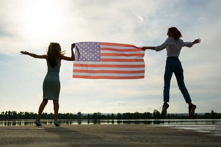 Two young friends women holding USA national flag jumping up together outdoors at sunset. Patriotic girls celebrating United States independence day.の写真素材