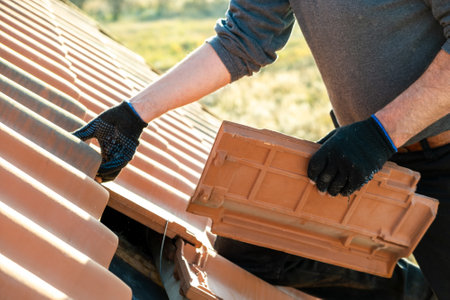 Closeup of worker hands installing yellow ceramic roofing tiles mounted on wooden boards covering residential building roof under construction.の写真素材
