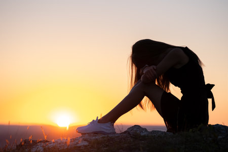 Young depressed woman in black short summer dress sitting on a mountain hill thinking outdoors at sunset. Lonely female contemplating in warm evening in nature.の写真素材