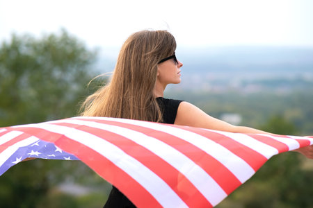 Young happy woman with long hair holding waving on wind american national flag on her sholders resting outdoors enjoying warm summer day.の写真素材