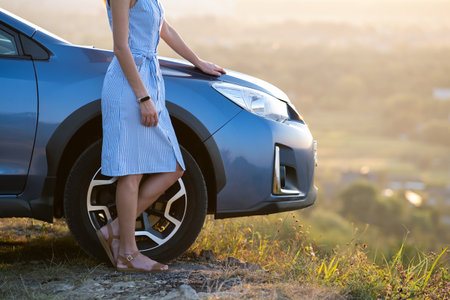 Closeup of young woman driver in blue dress leaning on her car enjoying warm summer day. Traveling and vacation concept.の写真素材