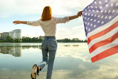 Red haired girl holding USA national flag in her hands. Positive young woman celebrating United States independence day.の写真素材