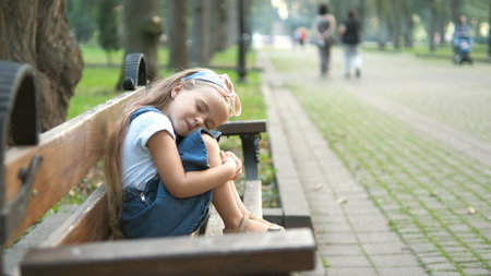 Small tired child girl sitting on a bench with closed eyes resting in summer park.の写真素材