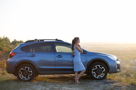 Happy woman driver in blue summer dress enjoying warm evening near her car. Travel and vacations...