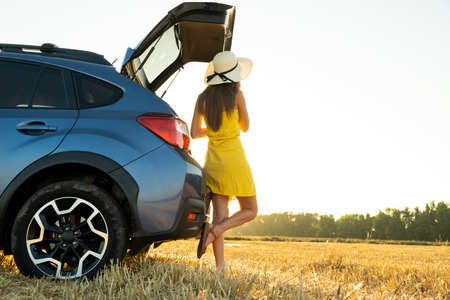 Young woman driver in yellow summer dress and straw hat standing near a blue car enjoying warm summer day at sunset.の写真素材