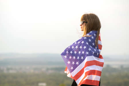 Young happy woman with long hair holding waving on wind american national flag on her sholders...