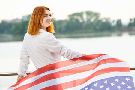 Happy red haired girl with USA national flag on her shoulders. Positive young woman celebrating United States independence day.の写真素材
