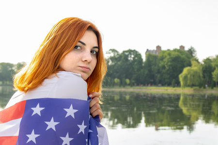 Portrait of sad red haired girl with USA national flag on her shoulders. Young woman celebrating United States independence day.の写真素材