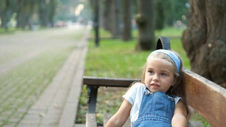 Small happy child girl sitting on a bench resting in summer park.