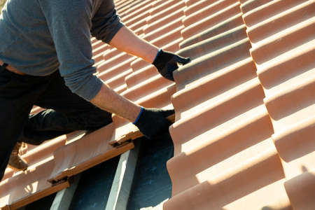 Closeup of worker hands installing yellow ceramic roofing tiles mounted on wooden boards covering residential building roof under construction.の写真素材