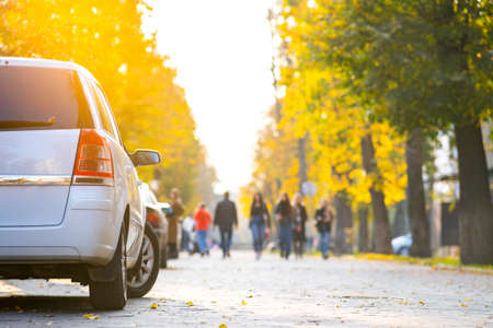 Cars parked in a row on a city street side on bright autumn day with blurred people walking on pedestrian zone.の写真素材