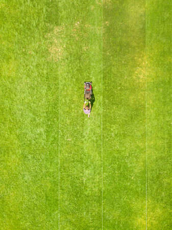 Aerial view of small figure of man worker trimming green grass with mowing mashine on football stadium field in summer.の写真素材