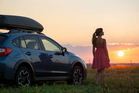 Dark silhouette of lonely woman relaxing near her car on grassy meadow enjoying view of colorful...
