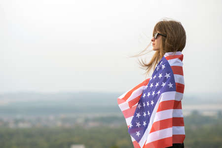 Young happy american woman with long hair holding waving on wind USA national flag on her sholders relaxing outdoors enjoying warm summer day.の写真素材