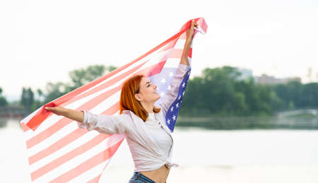Portrait of happy smiling red haired girl holding USA national flag up in her hands. Positive young woman celebrating United States independence day.の写真素材