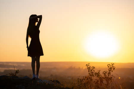 Dark silhouette of a young woman standing on a stone enjoying sunset view outdoors in summer.の写真素材