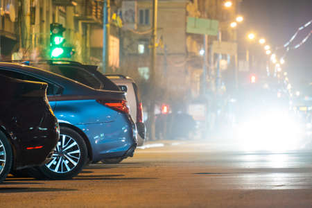 Close up of parked cars on roadside at night with blurred view of traffic lights of moving vehicles on city street.の写真素材